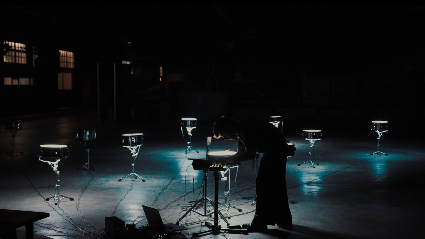 Eine Person steht in einem dunklen Raum zwischen mehreren beleuchteten Snare Drums und bedient elektronische Geräte für eine Klangperfomance