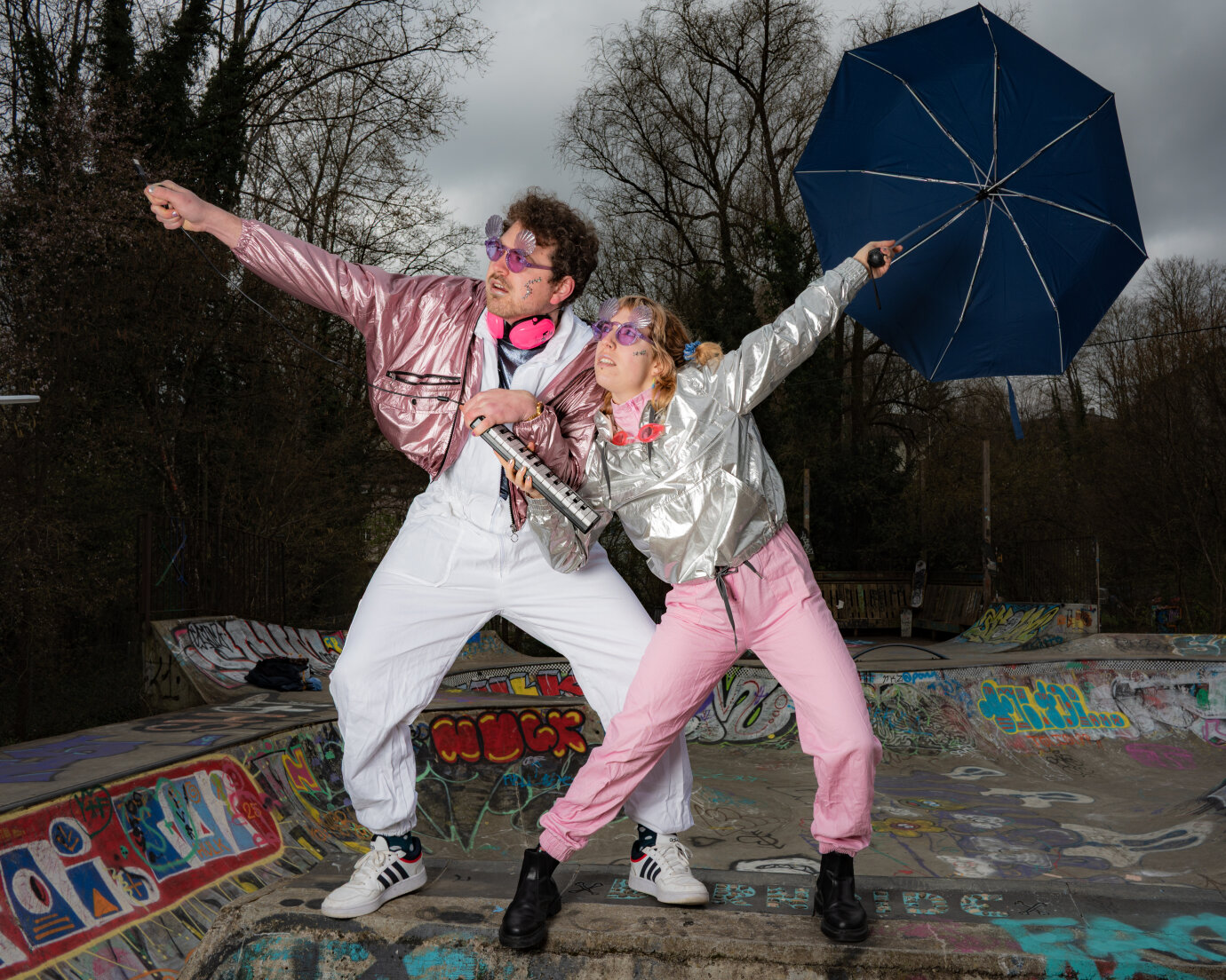 Mann und Frau in silbern und rosa glänzenden Jacken bewegen sich wie Disco-Tänzer*innen mit einer kleinen elektronischen Klaviatur und einem geöffneten Regenschirm in einem Skatepark.