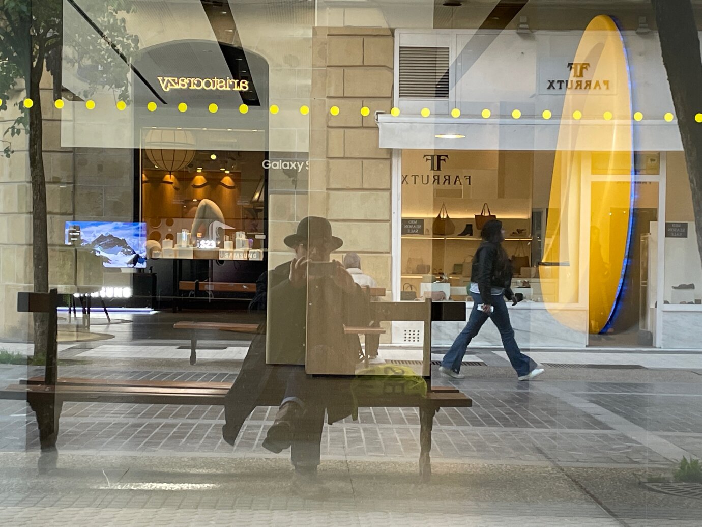 James Dillon sitzt auf einer Bank und fotografiert seine Spiegelung im Schaufenster eines Geschäfts mit dem Handy